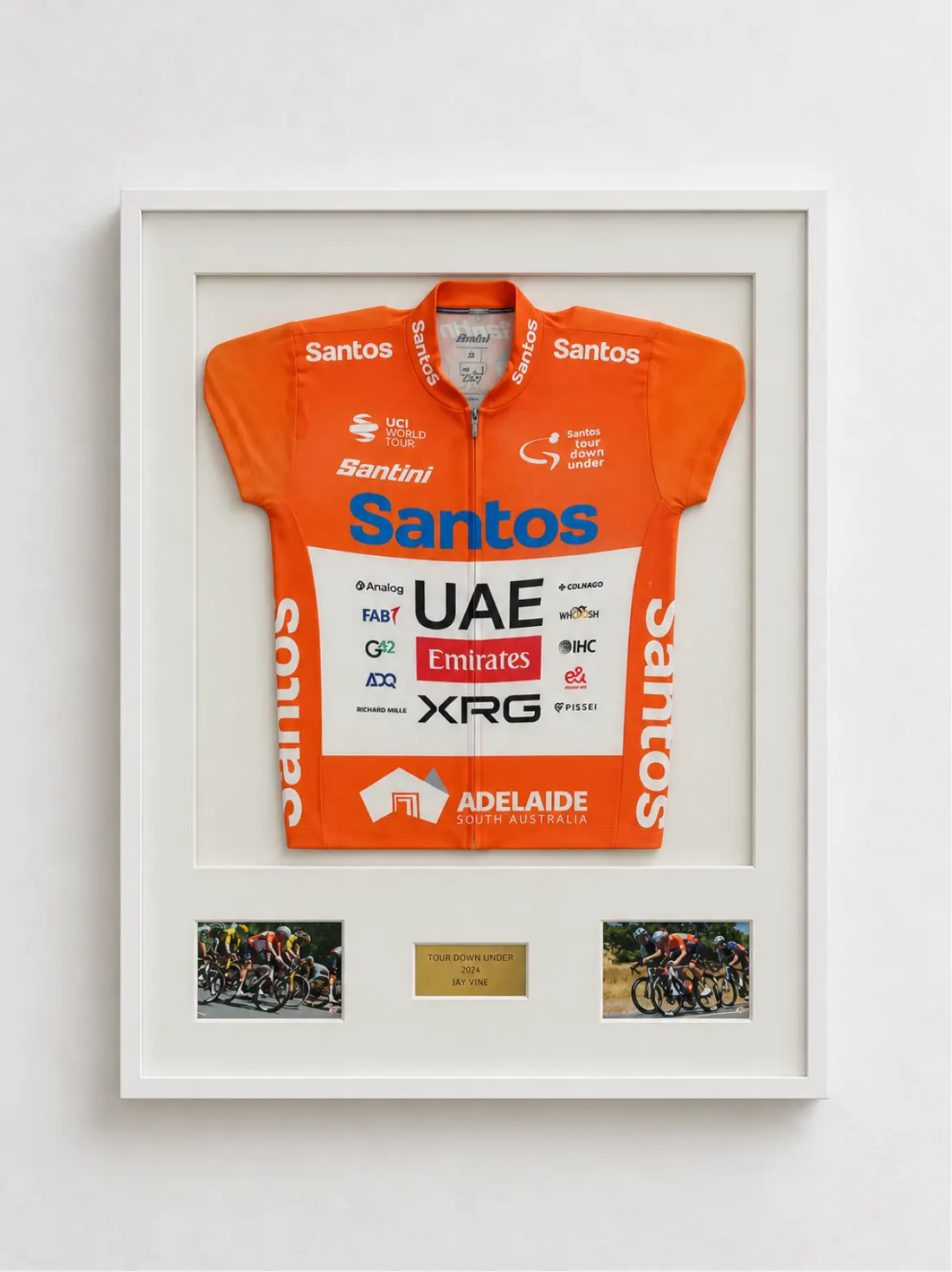Samarreta de ciclisme taronja amb patrocinadors, una placa i dues fotografies, en un marc blanc de caixa.